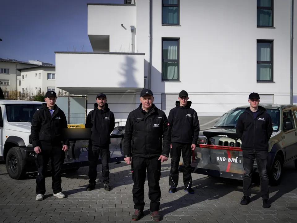 MDZ-Team aus fünf Mitarbeitern vor einem modernen weißen Mehrfamilienhaus, eingerahmt von zwei Winterdienst-Fahrzeugen mit Schneeschildern.